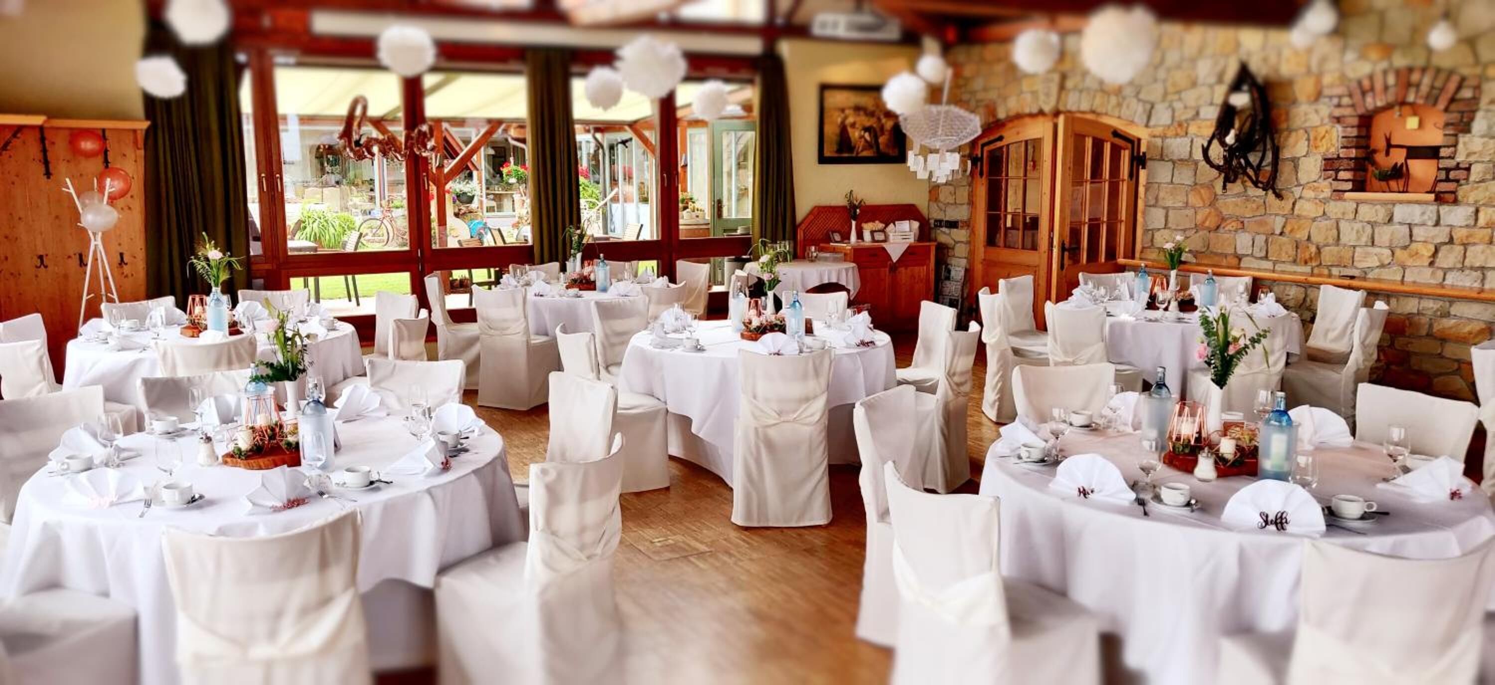 Une salle de banquet avec des tables rondes dressées pour un événement formel, agrémentée de nappes et de chaises blanches, d’un mur en pierre et d’une porte en bois.