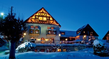 A large, multi-story building with a prominent gabled roof, illuminated windows, and snow-covered surroundings.