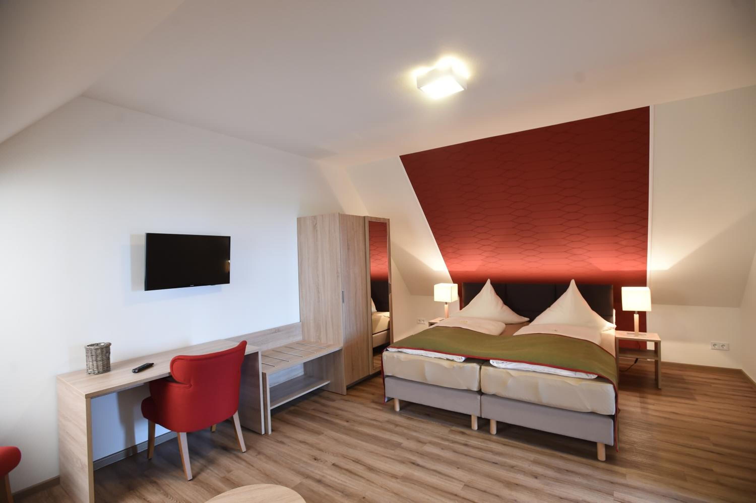 A modern bedroom with a desk, a bed with two pillows, a TV mounted on the wall, and a red accent wall.
