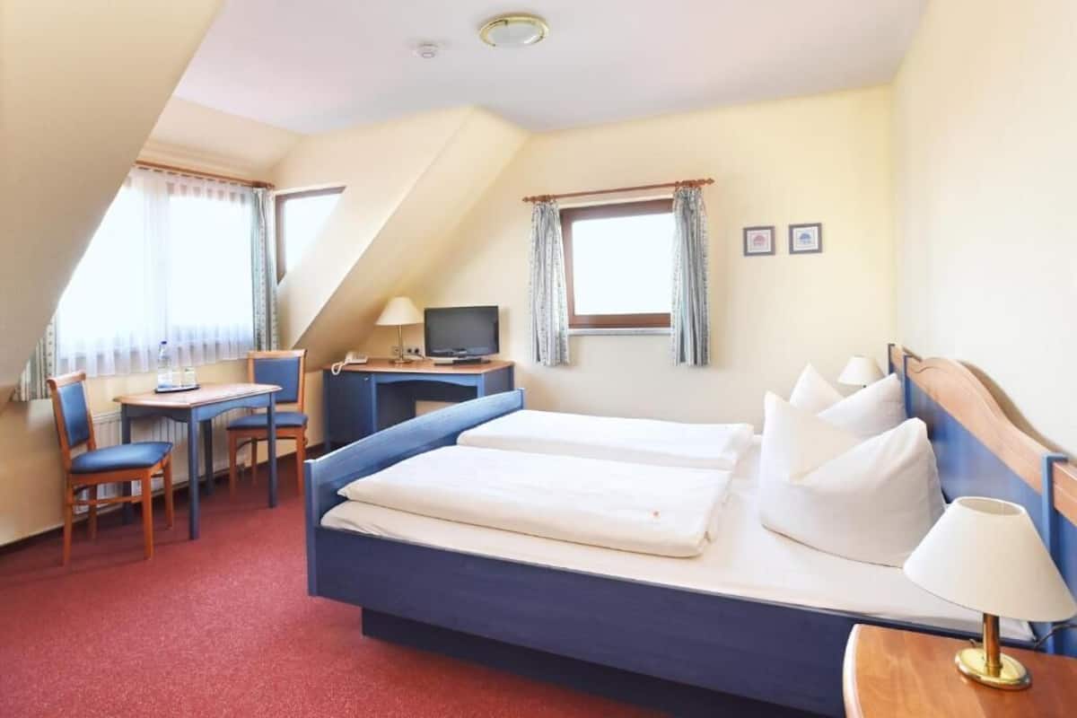 Quarto de hotel com cama, escrivaninha, cadeira, televisão e duas janelas com cortinas.