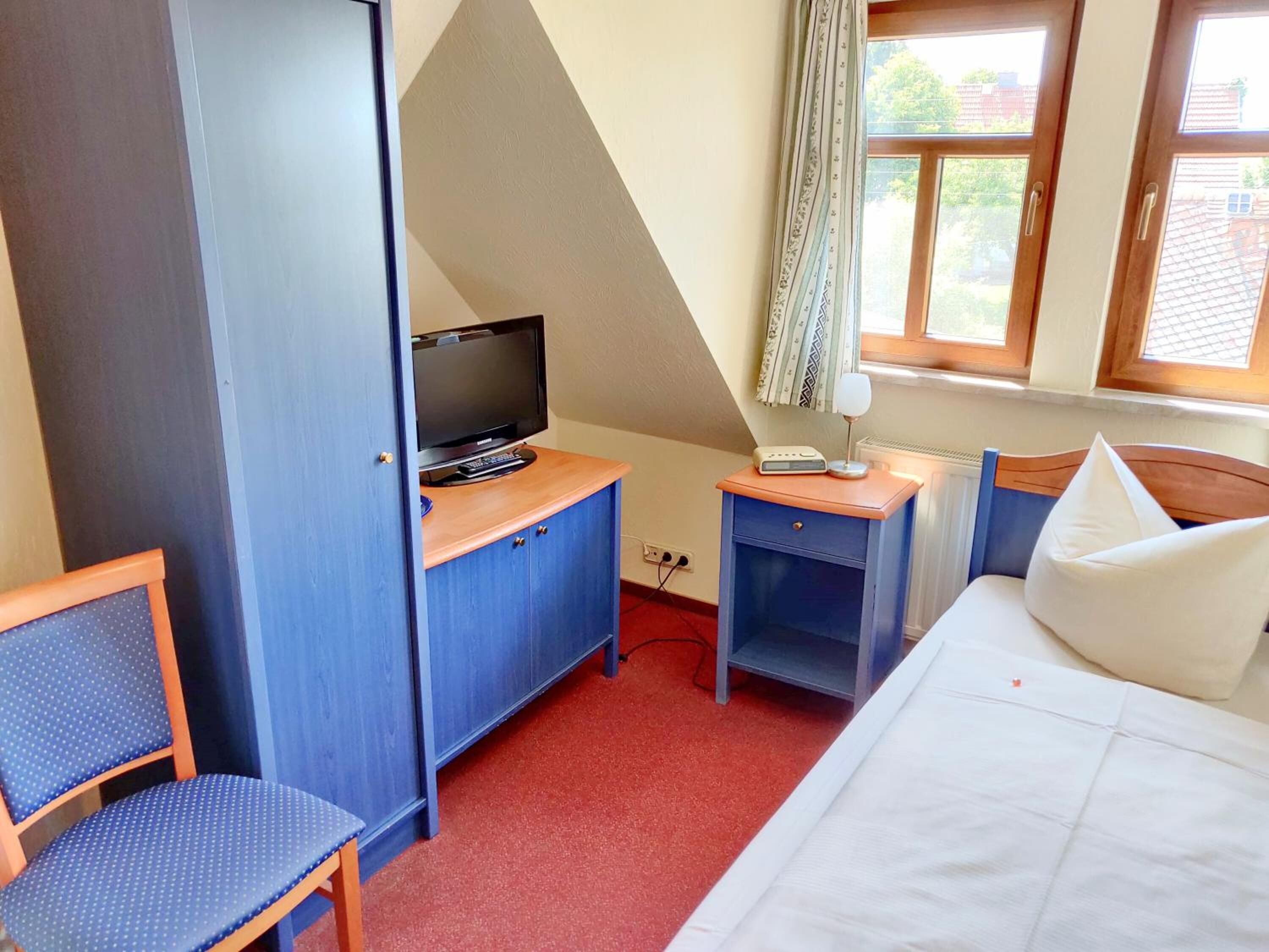Ein Schlafzimmer mit einem Bett, einem Schreibtisch mit Fernseher, einem Sessel und einem Fenster mit Vorhängen.