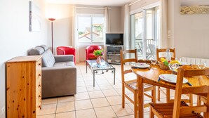 Ein modernes Wohnzimmer mit grauem Sofa, rotem Sessel und einem Holzesstisch, gedeckt f ür vier Personen. Ein Fernseher ist an der Wand befestigt, und durch die großen Fenster ist ein Balkon zu sehen.