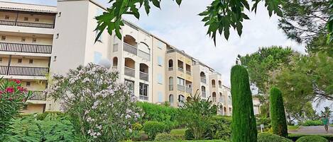 A multi-story residential building with balconies, surrounded by well-maintained gardens and trees.