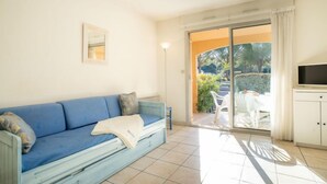 Ein Zimmer mit einem blauen Sofa, einem Fernseher auf einem Schrank und einer Glasschiebetür, die zu einem Außenbereich mit Pflanzen und einem Tisch führt.