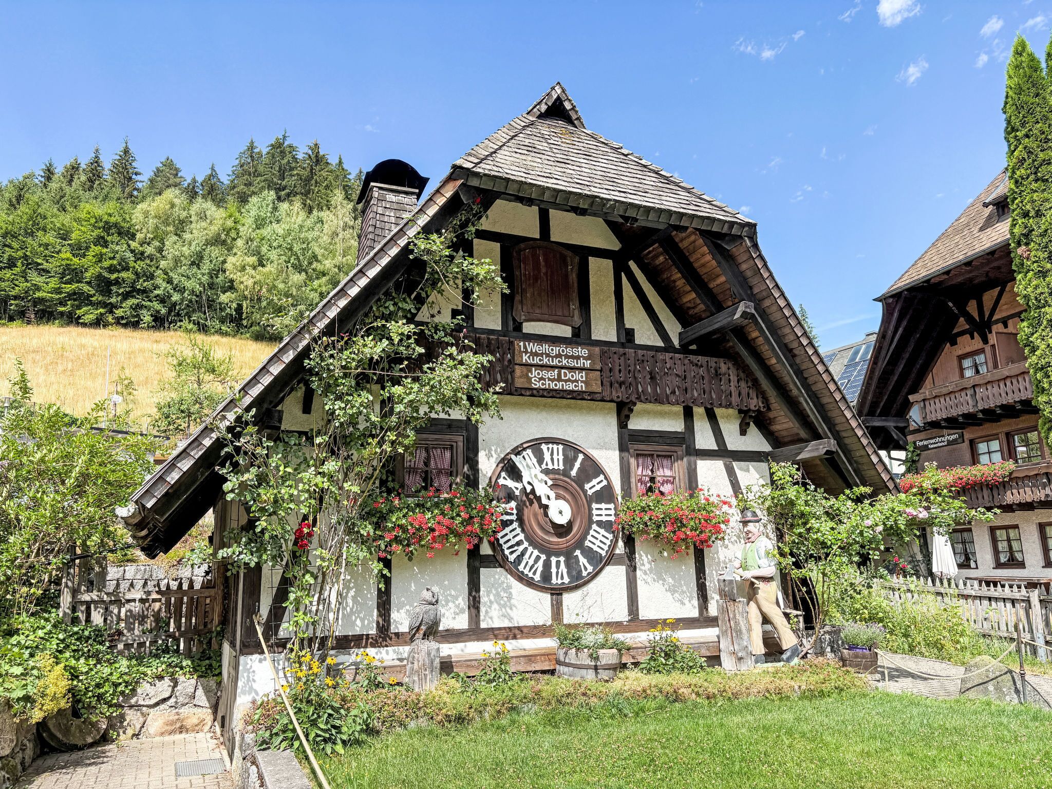A traditional half-timbered building with a large outdoor clock, surrounded by greenery and a clear sky.