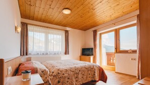 A bedroom with a wooden ceiling, a bed with floral bedding, a bedside table with a candle, a television on a wooden cabinet, and a view of mountains through a large window. - Voithofer by Interhome (Königsleiten)