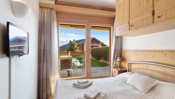 A hotel room with a bed, a TV, a balcony with a view of the mountains, and wooden furniture.