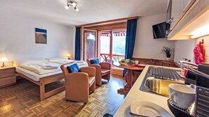 A hotel room with a bed, two chairs, a dining table with a bowl of fruit, a sink, and a television mounted on the wall. - Rose Studio Y by Interhome (Wengen)