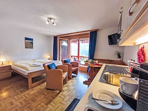 A hotel room with a bed, two chairs, a dining table with a bowl of fruit, a sink, and a television mounted on the wall. - Rose Studio Y by Interhome (Wengen)