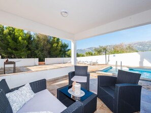 A covered outdoor area with a pool, wicker furniture, and a view of trees and hills.