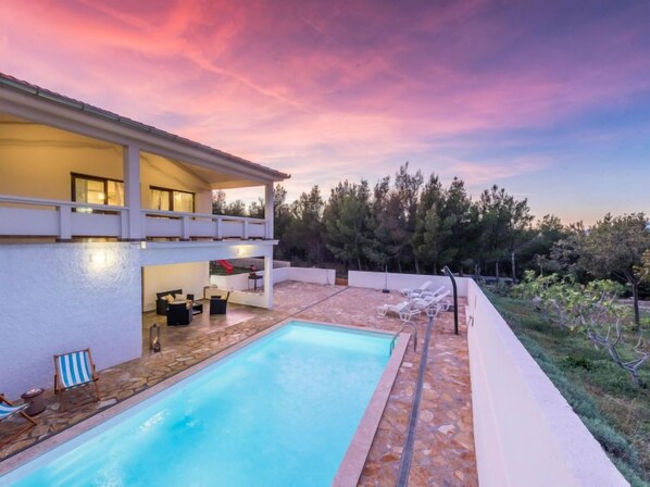 A two-story house with a swimming pool, outdoor seating, and a view of trees and hills.