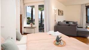 A hotel room with a bed, a sofa, a floor lamp, and a view of palm trees and mountains through the windows. - Suite Classic by Interhome (Ascona)