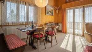 Un coin repas chaleureux avec des murs en bois, une grande fenêtre ornée de rideaux en dentelle, un luminaire suspendu et une table dressée pour quatre personnes.