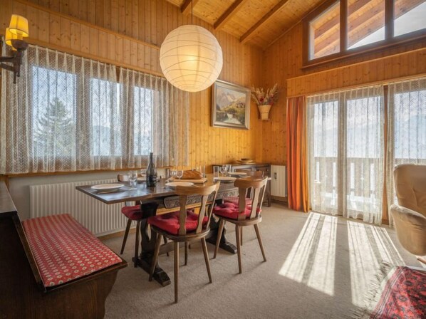 A cozy dining area with wooden walls, a large window with lace curtains, a hanging light fixture, and a table set for four.