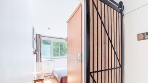 Ein Zimmer mit Holztür, einem Bett, einem kleinen Tisch und einem Fenster mit Blick auf Bäume.