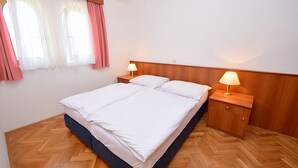 A double bed with two bedside tables, each with a lamp, in a room with wooden flooring and a window with curtains. - Meridiana by Interhome (Orebić)