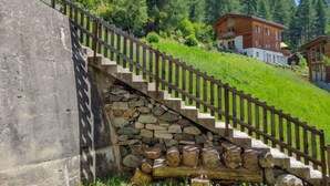 Un escalier en bois, adossé à un mur de pierre, et un banc en tronc d’arbre, qui mènent à une colline parsemée de maisons et d’arbres.