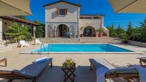 A poolside area with lounge chairs, a table with a drink, and a stone building with arched windows. - Histria 1 by Interhome (Pula/Vodnjan)