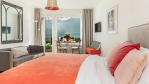 A bedroom with a large bed, a sofa, a dining table with chairs, a TV, and a view of the outdoors. - Double Room Modern by Interhome (Ascona)