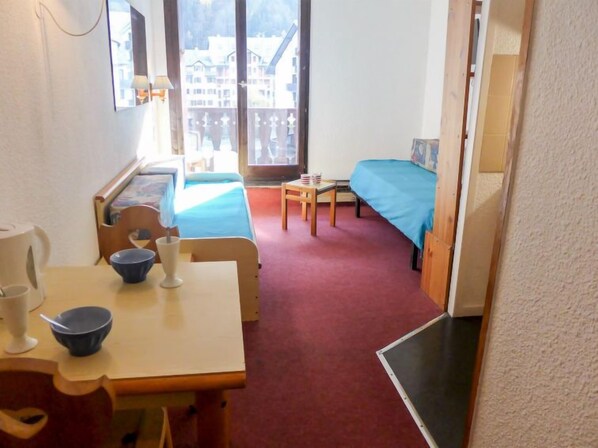 Una habitación pequeña y acogedora con una cama, una mesa con un tazón y una taza, y una ventana con vistas a edificios.