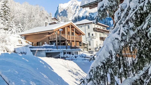Eine Holzhütte mit Balkon, umgeben von schneebedeckten Bäumen und Bergen im Hintergrund.