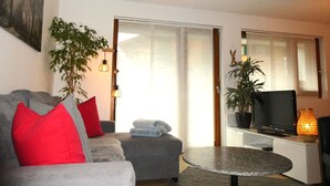 Ein modernes Wohnzimmer mit einem grauen Sofa, roten und grauen Kissen, einem kleinen runden Tisch, einem Fernseher und einer Topfpflanze.
