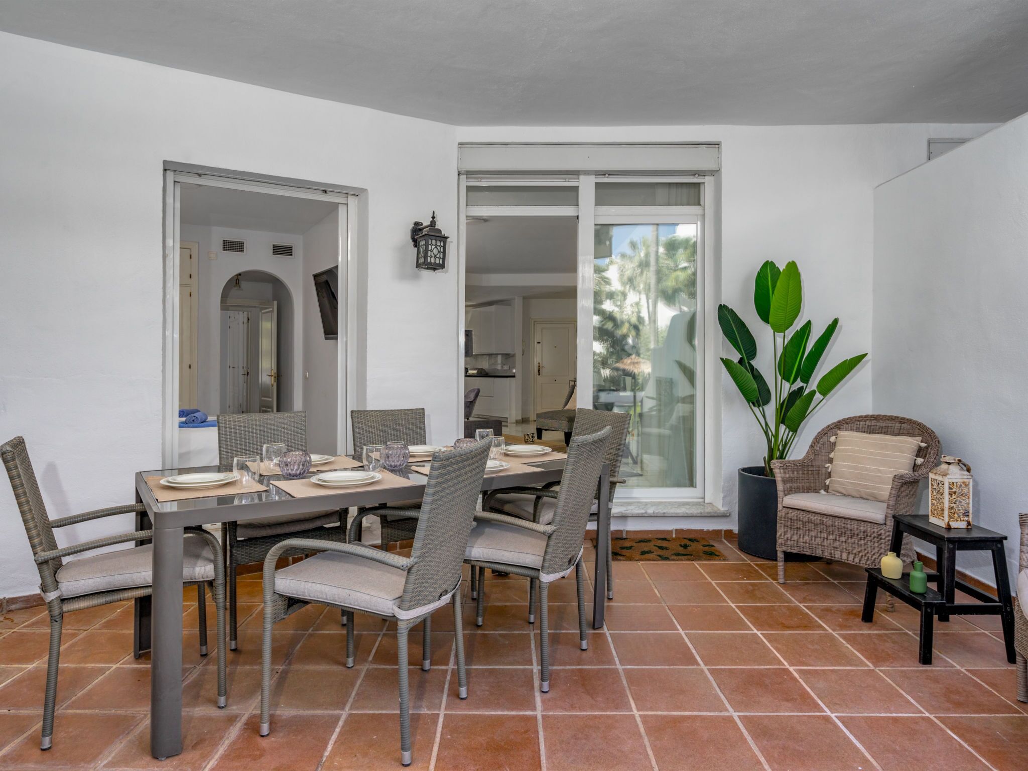 A dining area with a table set for six, surrounded by wicker chairs, a potted plant, and a wicker chair with cushions.