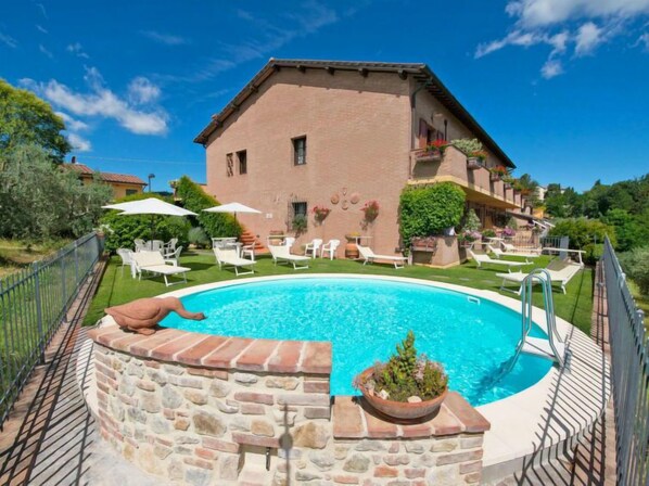 Vue sur une piscine aux murs de pierre, bordée de chaises longues et de parasols, avec un bâtiment en briques doté d’un balcon, et une végétation luxuriante en arrière-plan.