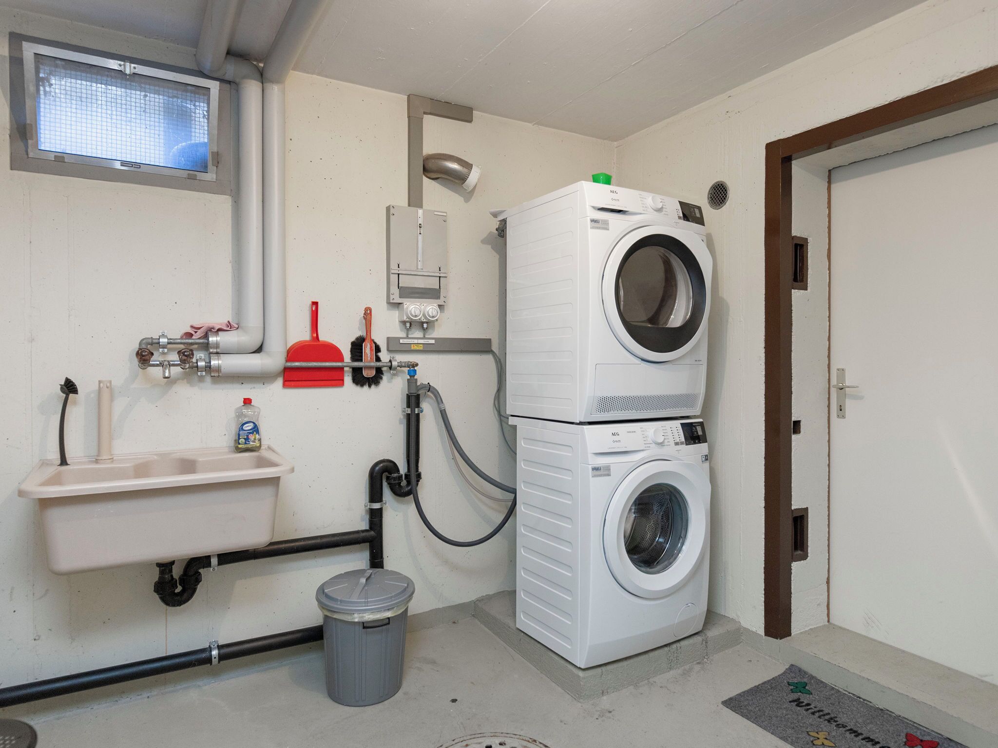 A laundry room with two stacked washing machines, a sink, a trash bin, and a window.