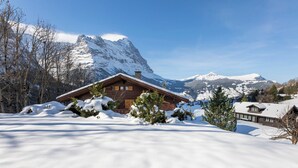 A wooden chalet with a sloped roof, surrounded by snow-covered mountains and trees.
