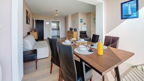 Een eethoek met een houten tafel, zwarte stoelen en een gele fles. Achterin staat een bank en een fauteuil.