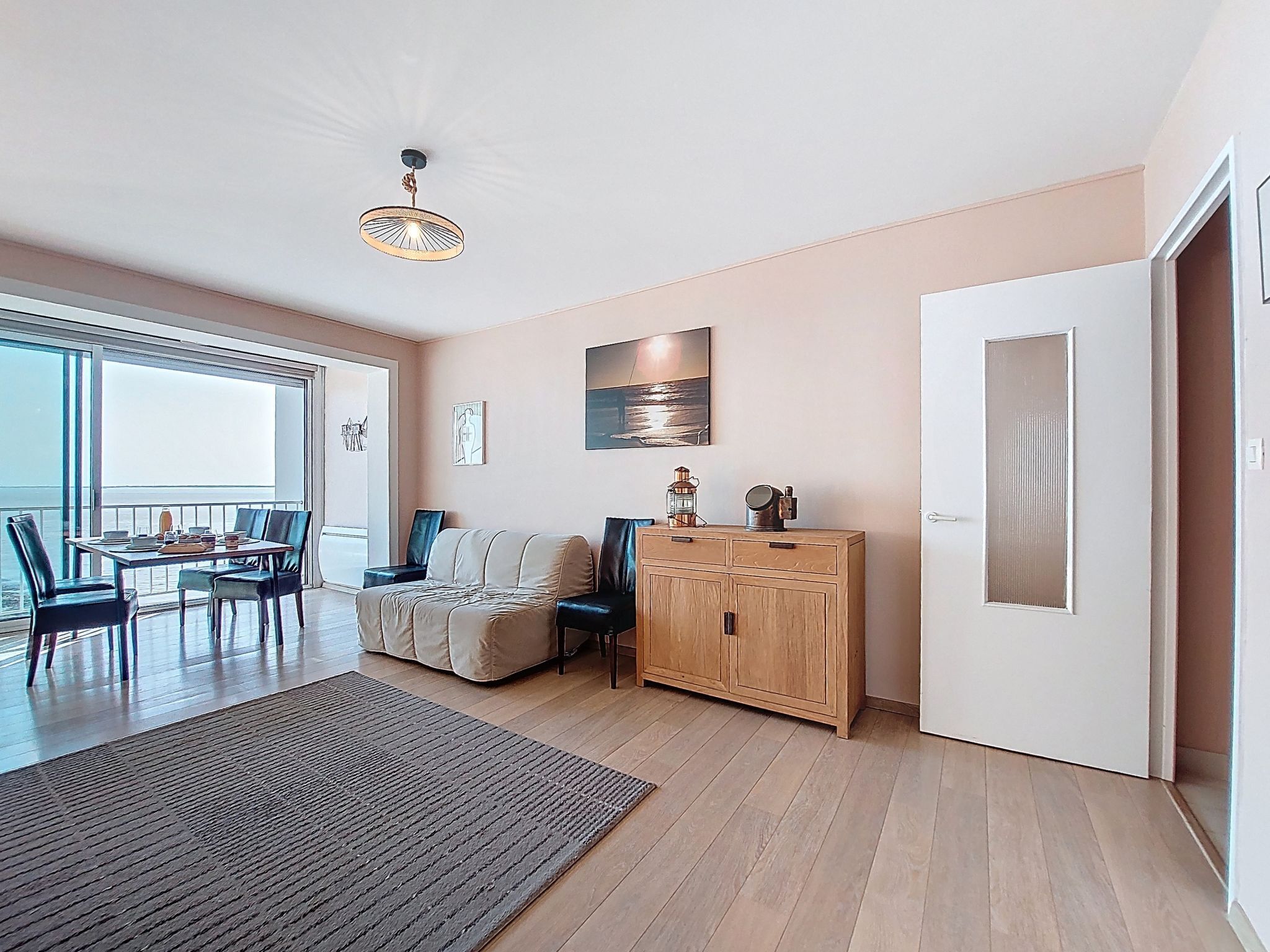 A spacious living area with a large window, a dining table, a sofa, a cabinet, and a door.