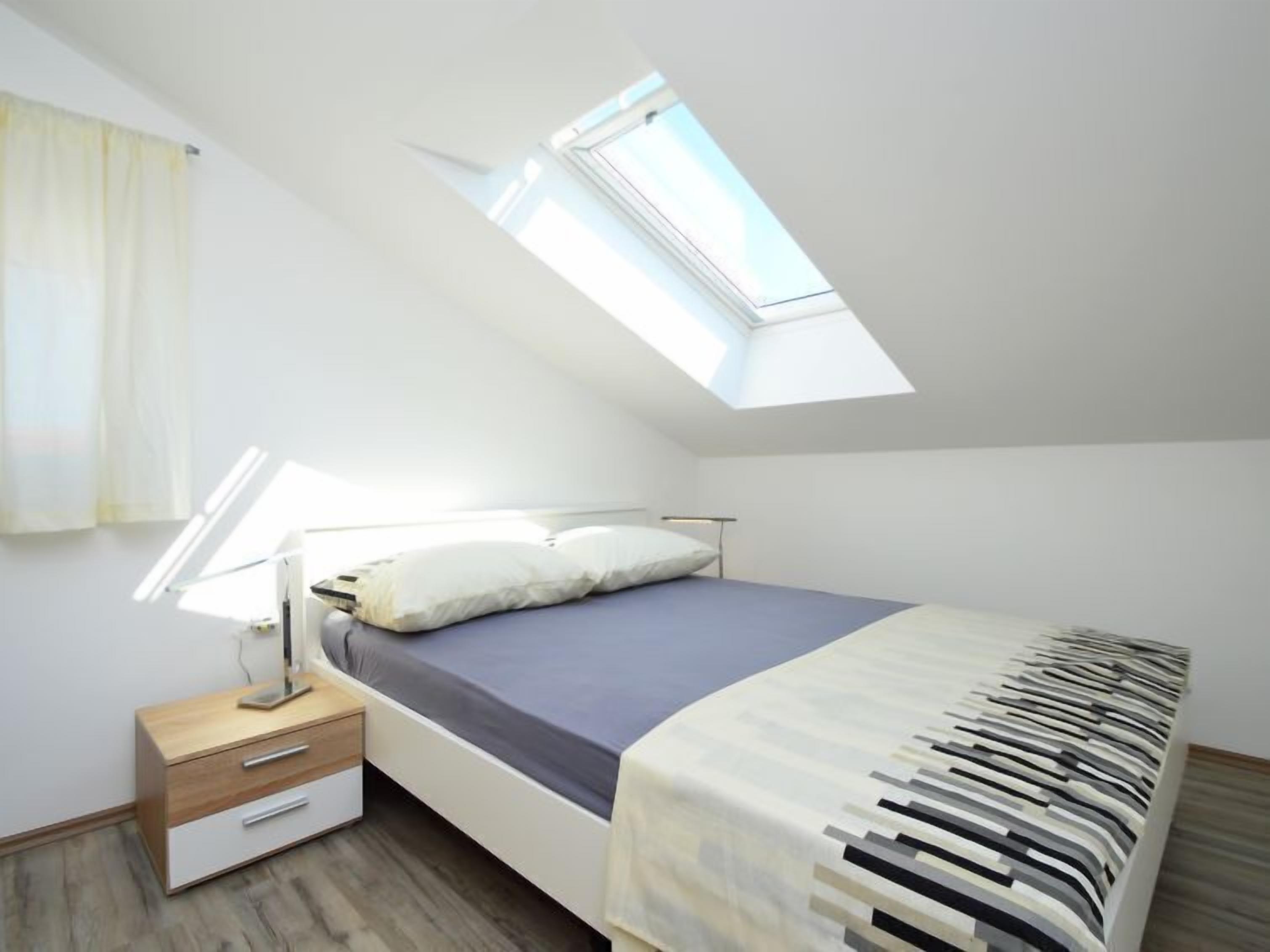 Un dormitorio con tragaluz, una cama con manta estampada, una mesita de noche con lámpara y un mueble auxiliar de madera.