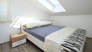 Une chambre avec un puits de lumière, un lit recouvert d’une couverture à motifs, une table de chevet avec une lampe et une commode en bois.