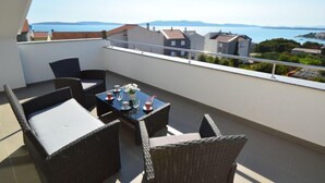 Un balcon agrémenté de meubles en osier et d’une table préparée pour deux personnes, offrant une vue sur la côte, avec des bâtiments et l’eau.