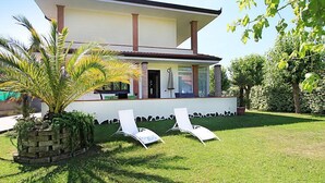 Une maison à deux étages avec un porche abrité, un jardin en briques agrémenté de plantes vertes et deux chaises longues blanches sur une pelouse bien entretenue.