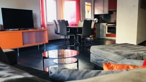Een moderne woonkamer met een bank, een glazen salontafel en een televisie op een kast. Er is een eethoek met stoelen en een keuken op de achtergrond.