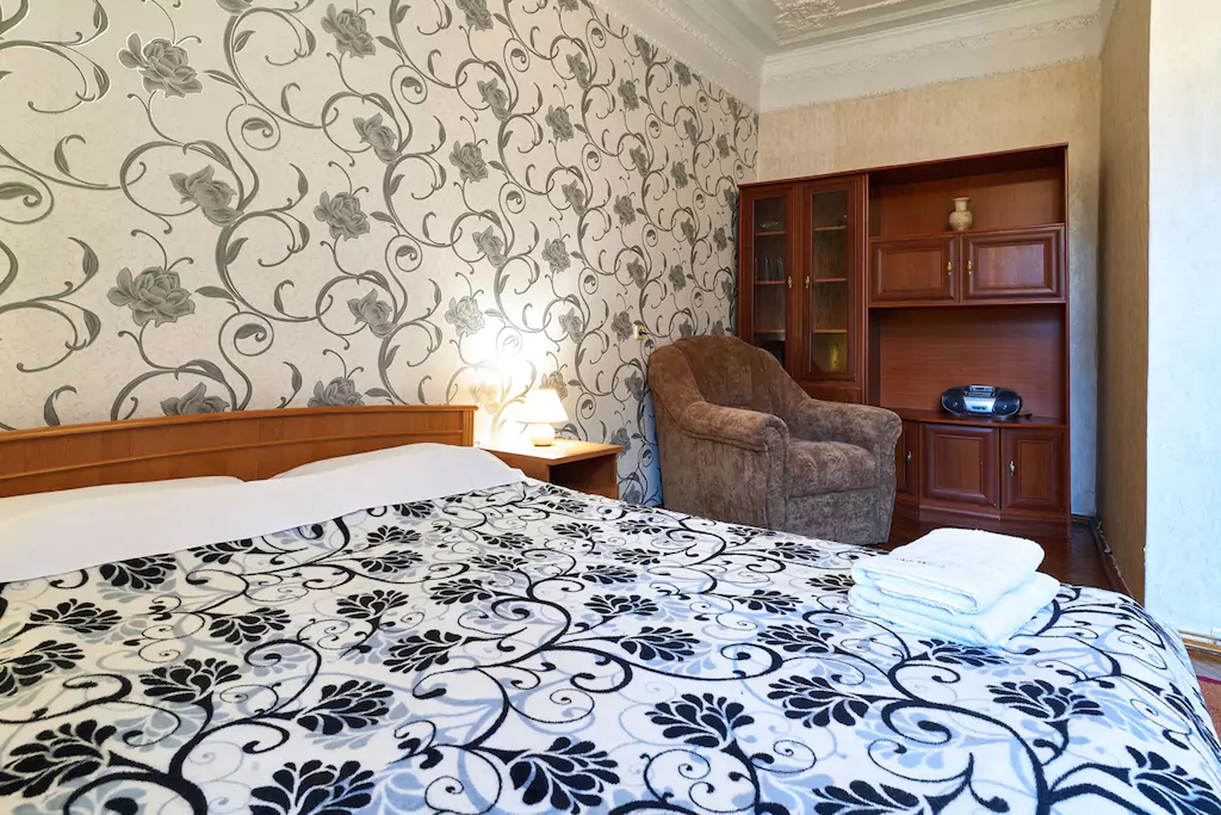 A hotel room with a large bed, a wooden nightstand, a lamp, a brown armchair, and a wooden cabinet with glass doors.