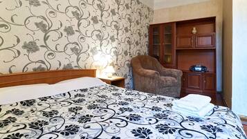 A hotel room with a large bed, a wooden nightstand, a lamp, a brown armchair, and a wooden cabinet with glass doors.