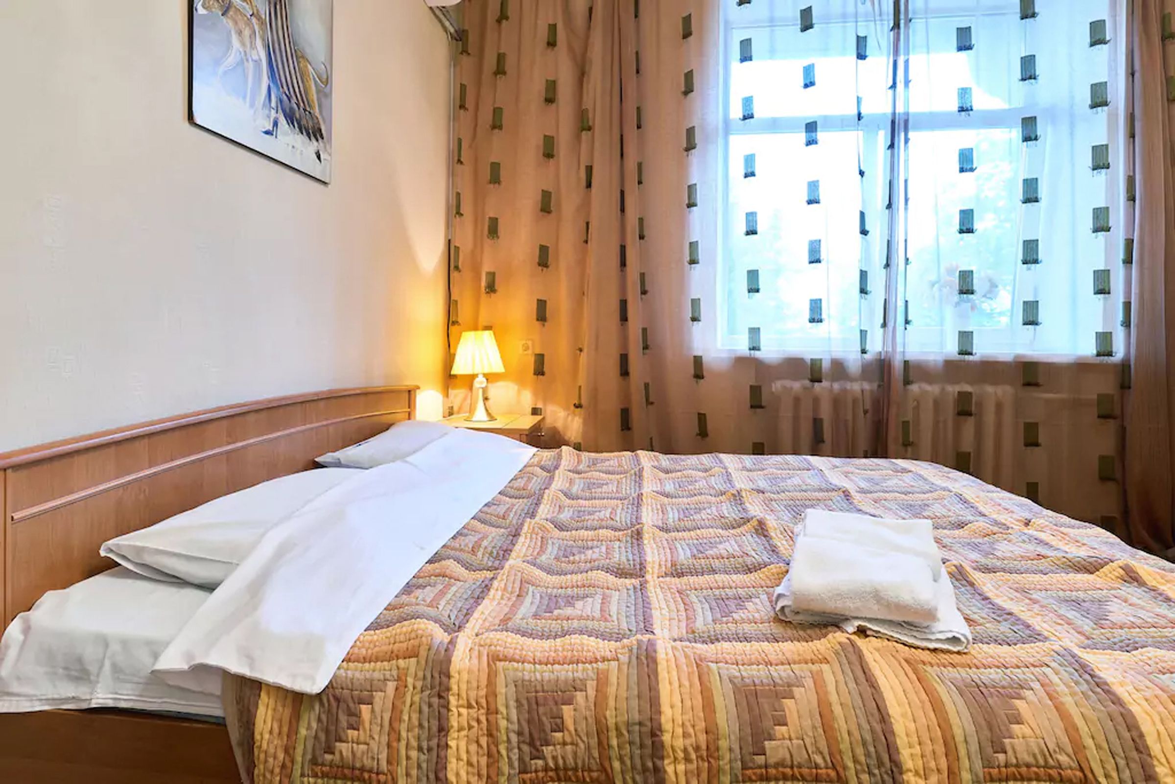 Quarto de hotel com cama bem arrumada, abajur na mesa de cabeceira e janela com cortinas.