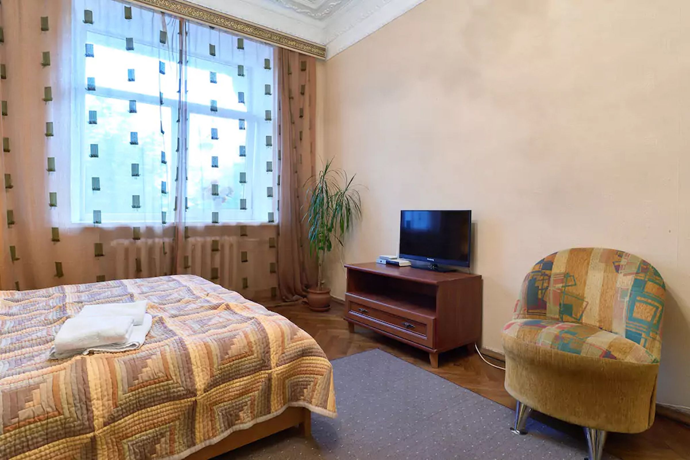 A hotel room with a bed, a television on a wooden stand, a chair, and a potted plant.