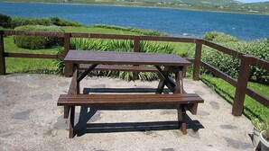 Et picnicbord med træbænk, omgivet af en stenbelagt plads og et træhegn, med udsigt over et vandareal og grønne bakker.