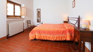 Un dormitorio con cama, una silla, una mesita de noche, una lámpara, una ventana y un radiador.