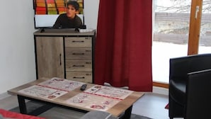 A room with a TV on a wooden cabinet, a table with magazines, and a window with red curtains. - 300m from ski slopes, floor -1, view mountain, parking, tv, ski locker, 36m², Valfréjus (Valfréjus)