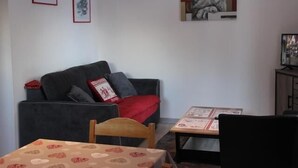 A living room with a grey sofa, a wooden chair, a small table with a patterned tablecloth, and framed pictures on the wall. - 300m from ski slopes, floor -1, view mountain, parking, tv, ski locker, 36m², Valfréjus (Valfréjus)