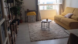 Een woonkamer met een bank, een rond bijzettafeltje, een boekenkast, een potplant en een raam met gordijnen.