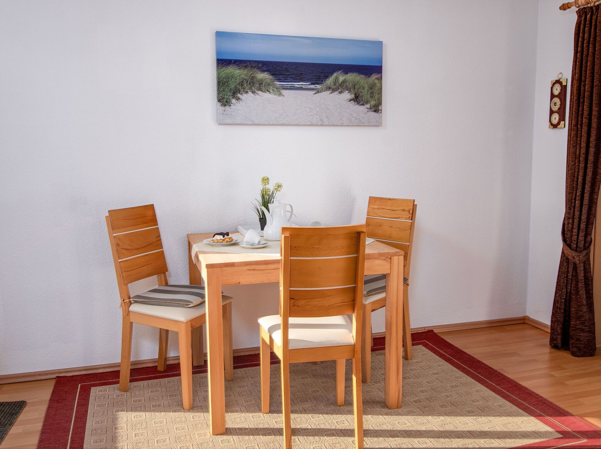 Un coin repas avec une table et des chaises, un tableau représentant une scène de plage et une horloge au mur.