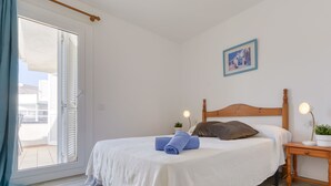 Ein ordentlich gemachtes Bett mit weißer Bettdecke, einem blauen Handtuch und einem braunen Kopfkissen. Auf dem Nachttisch steht ein kleiner Topfpflanze, und an der Wand hängt ein Bilderrahmen.