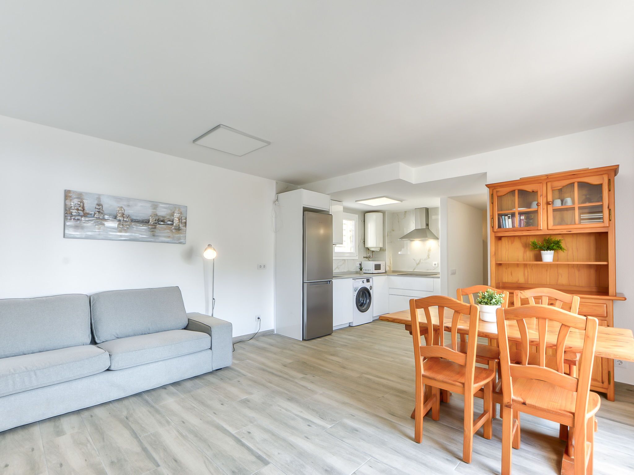 A modern living room with a grey sofa, wooden dining table with chairs, a kitchen with a washing machine, and a cabinet with glass doors.