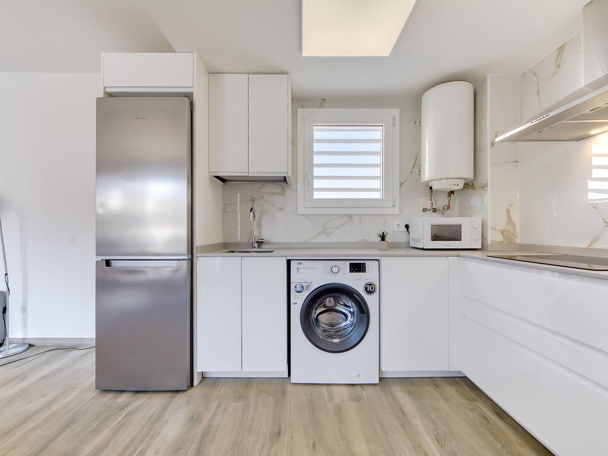 Una cocina moderna con un refrigerador de acero inoxidable, lavadora integrada, microondas y ventana con persianas.